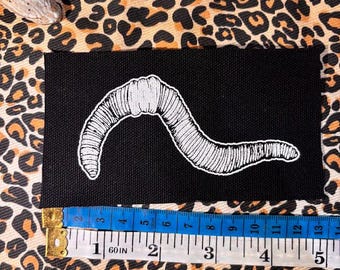May include: A rectangular black fabric patch featuring a white line-art illustration of an earthworm. A measuring tape is positioned along the bottom edge, showing the patch is approximately 13 centimetres wide and 5 inches in length.