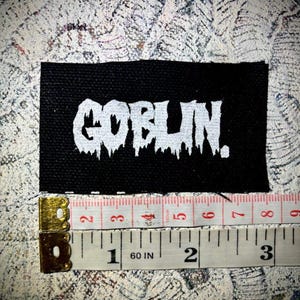 Könnte beinhalten: Schwarzer Stoff-Patch mit weißem Text, der "GOBLIN." lautet. Der Patch ist etwa 5 cm breit und 2,5 cm hoch.