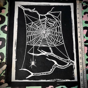 Op de afbeelding: Zwart en wit stoffen patch met een spinnenwebontwerp. Het web staat op een zwarte achtergrond, omlijst door een witte rand. Een spin hangt aan het web. De patch meet ongeveer 25 cm bij 20 cm.