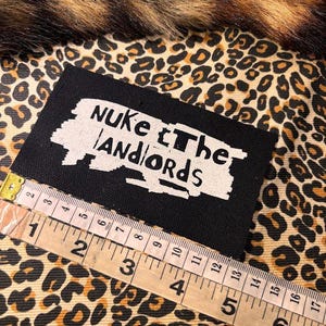 Peut inclure: Un &eacute;cusson rectangulaire noir avec l'inscription blanche "NUKE the LANDLORDS". Un m&egrave;tre ruban est plac&eacute; &agrave; c&ocirc;t&eacute; du patch. Le fond est un motif l&eacute;opard.