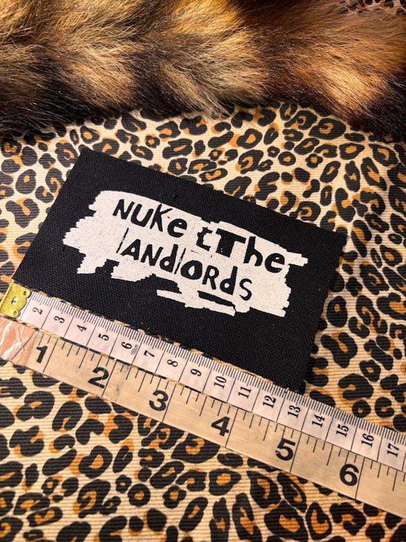 Peut inclure: Un &eacute;cusson rectangulaire noir avec l'inscription blanche "NUKE the LANDLORDS". Un m&egrave;tre ruban est plac&eacute; &agrave; c&ocirc;t&eacute; du patch. Le fond est un motif l&eacute;opard.