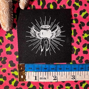 May include: A square black fabric patch featuring a white embroidered graphic of a winged tooth with a halo and radiating sunburst lines. A measuring tape is positioned at the bottom, showing the patch width is approximately 3 inches.