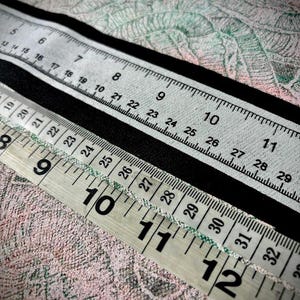 May include: A close-up of two white measuring tapes with black markings. The top tape is partially obscured by a black strip. The bottom tape shows numbers from 9 to 12, with markings for inches.