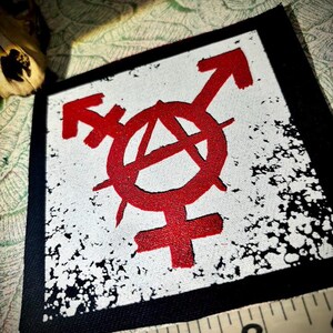 May include: A square fabric patch featuring a red anarchy symbol combined with gender symbols on a white background with black distressed edges. A measuring tape is visible at the bottom, indicating the patch is approximately 3 inches wide.