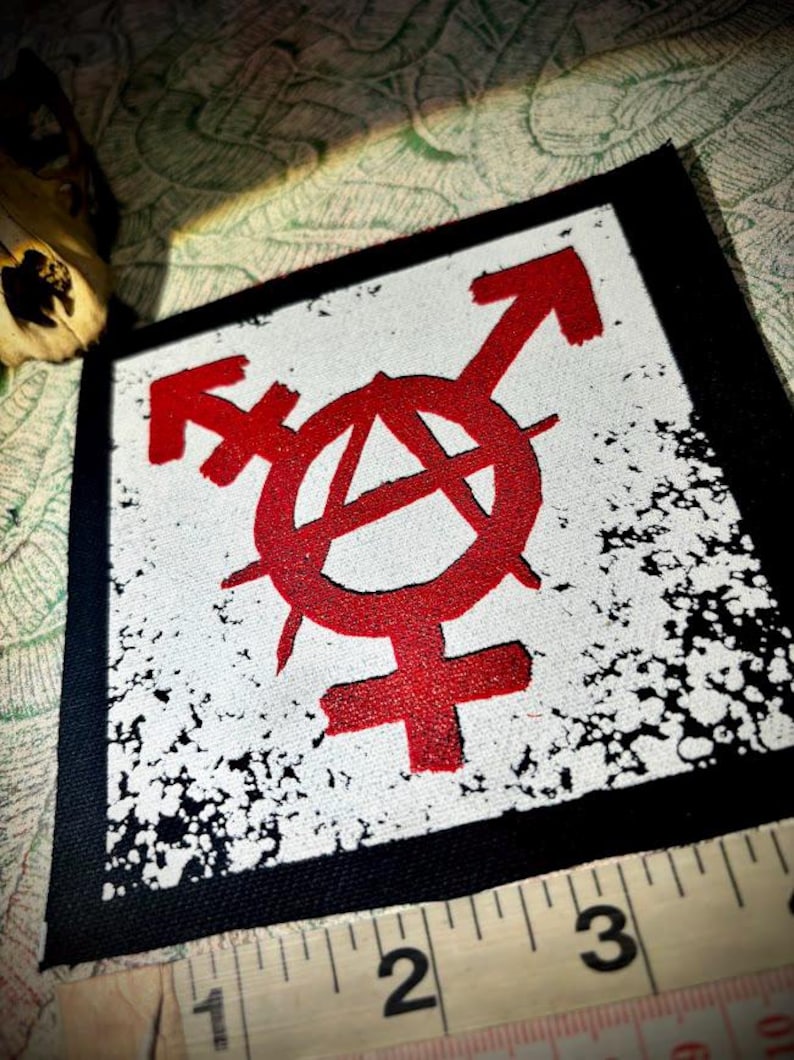 May include: A square fabric patch featuring a red anarchy symbol combined with gender symbols on a white background with black distressed edges. A measuring tape is visible at the bottom, indicating the patch is approximately 3 inches wide.
