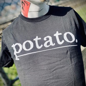 Op de afbeelding: Een zwart T-shirt met een witte afbeelding waarop "potato." staat. De afbeelding is in een gestileerd lettertype.