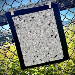 Peut inclure: Une œuvre d'art encadrée en noir avec un motif abstrait détaillé en blanc sur fond noir. L'œuvre est maintenue par des clips sur une clôture en treillis métallique, avec un ciel bleu visible en arrière-plan. Le motif est composé de nombreuses formes interconnectées en forme de ver.