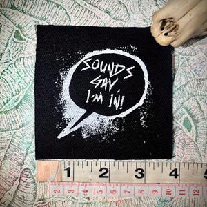 Könnte beinhalten: Schwarzer quadratischer Aufnäher mit dem Text "SOUNDS GAY I'M IN!" in einer weißen Sprechblase. Der Aufnäher ist etwa 10 cm breit. Der Aufnäher befindet sich auf einer gemusterten Oberfläche.