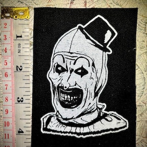 May include: Black and white embroidered patch of a creepy clown with a top hat. The clown has a wide, toothy grin and exaggerated features.