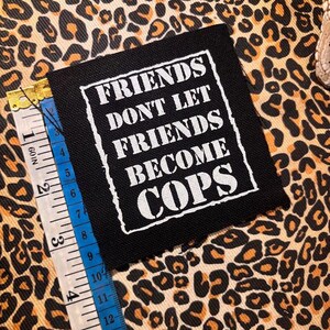 May include: A square black fabric patch with white embroidered text that reads "FRIENDS DONT LET FRIENDS BECOME COPS" inside a white border. A measuring tape is placed along the left edge, showing the patch is approximately 3.5 inches wide.