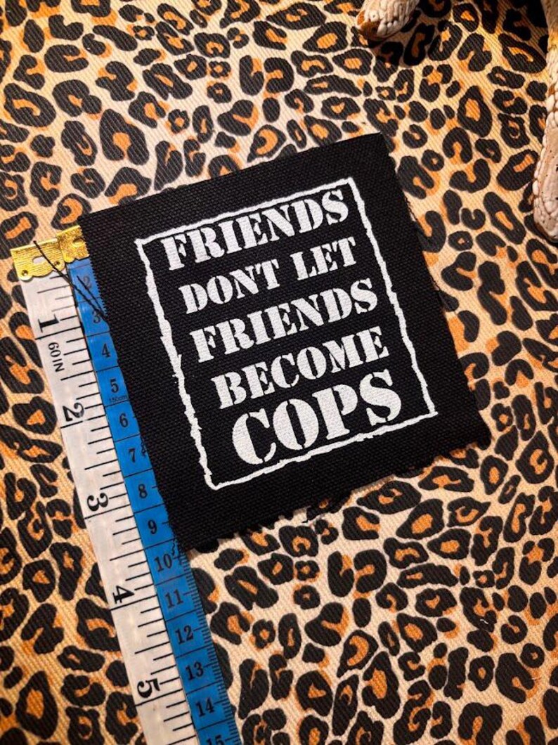 May include: A square black fabric patch with white embroidered text that reads "FRIENDS DONT LET FRIENDS BECOME COPS" inside a white border. A measuring tape is placed along the left edge, showing the patch is approximately 3.5 inches wide.