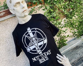 May include: Black t-shirt with a white pentagram design and the text "NOT TODAY JESUS." The shirt is displayed on a mannequin with a weathered appearance, set against a rustic wooden surface and a wall with climbing plants.