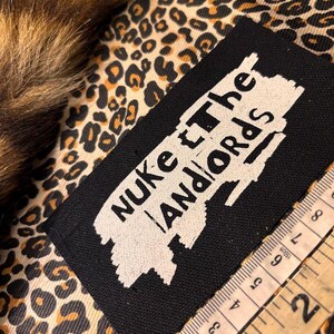 Peut inclure: &Eacute;cusson rectangulaire noir avec les mots "Nuke the Landlords" en blanc. L'&eacute;cusson est sur un tissu &agrave; imprim&eacute; l&eacute;opard avec un m&egrave;tre ruban en centim&egrave;tres. Une garniture en fourrure marron et beige est visible en haut &agrave; gauche.