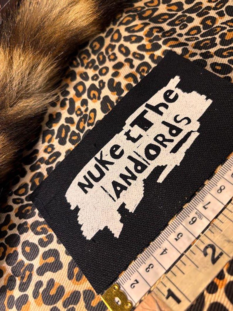 Peut inclure: &Eacute;cusson rectangulaire noir avec les mots "Nuke the Landlords" en blanc. L'&eacute;cusson est sur un tissu &agrave; imprim&eacute; l&eacute;opard avec un m&egrave;tre ruban en centim&egrave;tres. Une garniture en fourrure marron et beige est visible en haut &agrave; gauche.