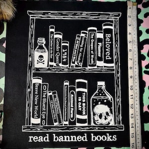 Könnte beinhalten: Schwarz-weiß-Illustration eines Bücherregals mit einem Schädel in einer Flasche. Das Bücherregal ist gefüllt mit Büchern mit Titeln wie "Beloved", "Flamer", "The Grapes of Wrath", "Gender Queer", "1984" und "The Catcher in the Rye". Der Text "read banned books" befindet sich am unteren Rand des Bildes.