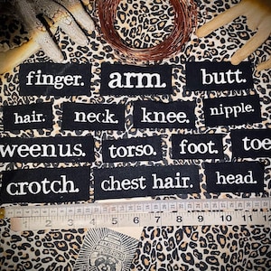 May include: Black and white fabric patches with various body part names printed on them, including finger, arm, butt, hair, neck, knee, nipple, weenus, torso, foot, toe, crotch, chest hair, and head.