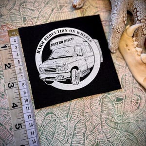 May include: A black square patch featuring a white van illustration and text. The text reads "HARM REDUCTION ON WHEELS" and "DISTRO DISCO". A measuring tape is positioned next to the patch, showing measurements in inches.