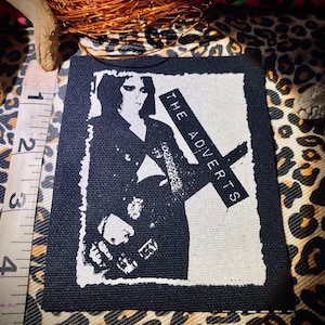 Könnte beinhalten: Schwarz-weißer Aufnäher mit dem Text "THE ADVERTS" auf einem Kreuz, der eine stilisierte Illustration einer Person zeigt. Der Aufnäher hat einen schwarzen Rand und ist wahrscheinlich aus Stoff.