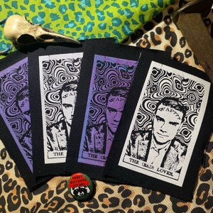 Pode incluir: Três patches de tecido preto com desenhos brancos e roxos. Cada patch apresenta um retrato de uma pessoa com o texto "WAP" acima de sua cabeça. O texto "THE (BAD) LOVER" está impresso na parte inferior do patch mais à direita.