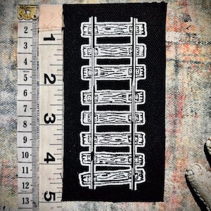Könnte beinhalten: Schwarzer Stoff-Patch mit einem weißen Umriss eines Gleises. Das Gleis besteht aus Holzschwellen und Metallschienen.