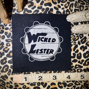 Pode incluir: Remendo de tecido preto com um design circular branco. O círculo tem um pandeiro branco com o texto "Wicked Lester" dentro.