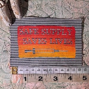 May include: A rectangular patch with the text "SAFE SUPPLY SAVES LIVES" in white letters on a red to orange gradient background. A syringe graphic is below the text. The patch is approximately 10 cm wide and has a striped border.