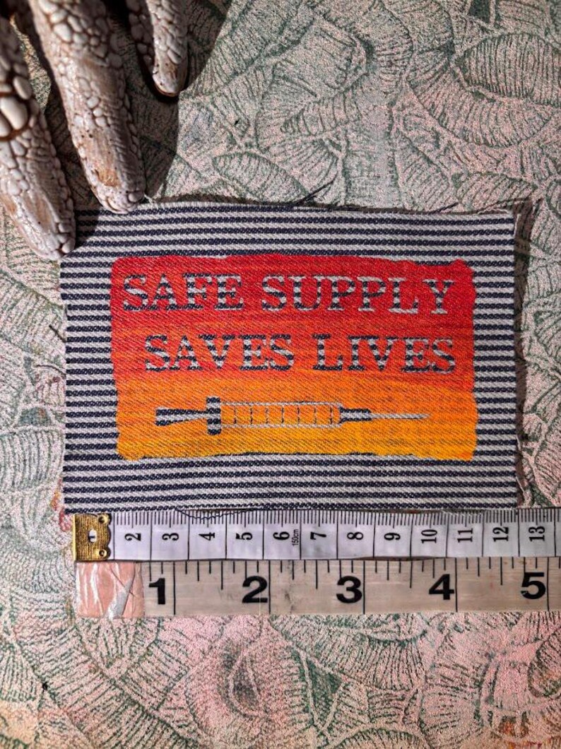 May include: A rectangular patch with the text "SAFE SUPPLY SAVES LIVES" in white letters on a red to orange gradient background. A syringe graphic is below the text. The patch is approximately 10 cm wide and has a striped border.