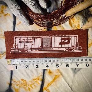 May include: A brown fabric patch with a white outline of a boxcar. The boxcar has the text "BURLINGTON NORTHERN BN 355145" on the side. The patch is approximately 8 inches long and 2 inches wide.