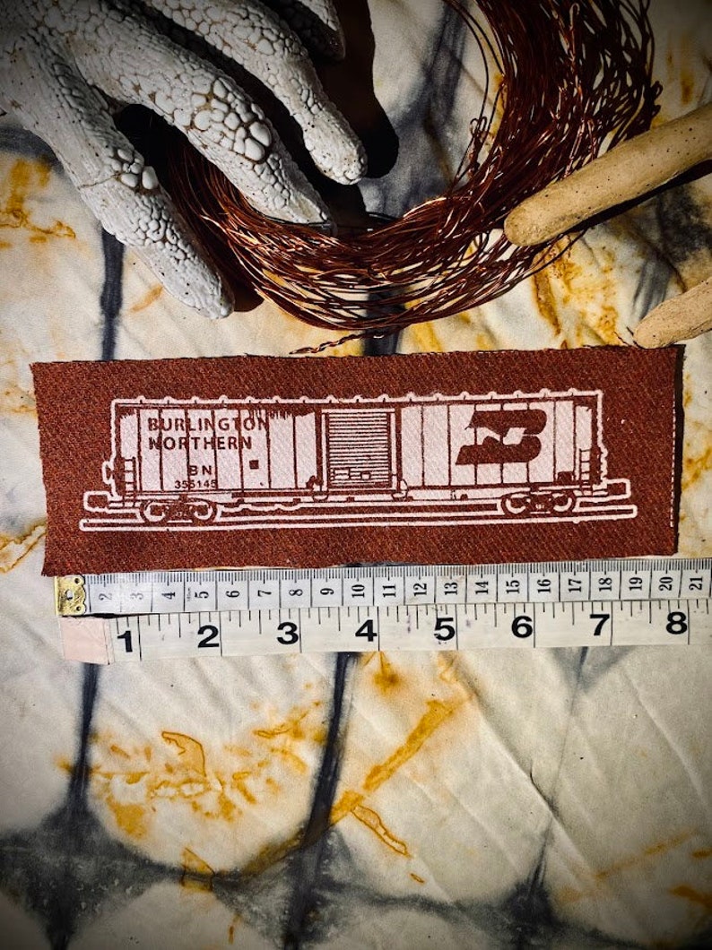 May include: A brown fabric patch with a white outline of a boxcar. The boxcar has the text "BURLINGTON NORTHERN BN 355145" on the side. The patch is approximately 8 inches long and 2 inches wide.