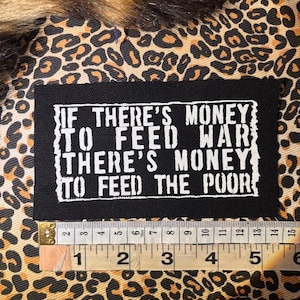 Pode incluir: Remendo retangular preto com texto branco: "IF THERE'S MONEY TO FEED WAR THERE'S MONEY TO FEED THE POOR." O remendo está em um fundo com estampa de leopardo. Uma régua mostra que o remendo tem aproximadamente 13 cm de largura.