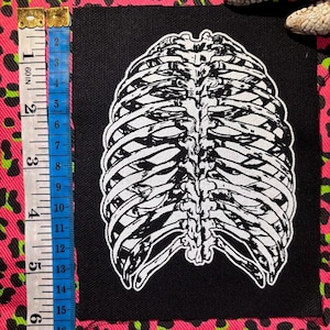 May include: A black fabric patch featuring a white screen-printed human rib cage illustration. A measuring tape placed alongside the patch indicates a height of approximately 7 inches, confirming the scale of the rectangular textile item.