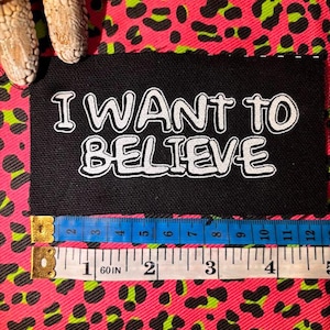 May include: A rectangular black embroidered patch with the white text I WANT TO BELIEVE. A measuring tape is positioned along the bottom edge, showing a width of approximately 4.5 inches. The patch is placed on a bright pink and green leopard print.