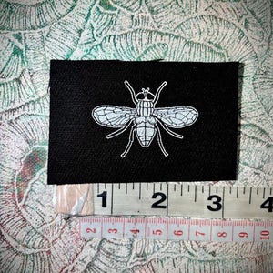 May include: A black rectangular patch with a white fly design. The patch is about 7.6 cm wide. The fly illustration has detailed wings, body segments, and legs. The patch is displayed on a patterned background.