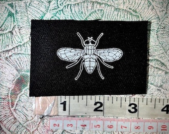 May include: A black rectangular patch featuring a white fly illustration. The patch is approximately 3 inches wide. The fly design includes detailed wings, body segments, and legs. The patch is placed on a patterned background.