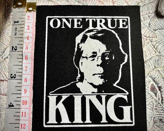 以下が含まれることがあります： 眼鏡をかけた人物の白いシルクスクリーン肖像画が描かれた長方形の黒い布製パッチ。上部に「ONE TRUE」、下部に「KING」の文字がある。横に置かれたメジャーは、高さが約14cmであることを示している。