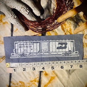 May include: A rectangular navy and white gingham fabric patch featuring a white embroidered outline of a Burlington Northern train car. A measuring tape is positioned along the bottom edge, showing a length of approximately 8 inches.