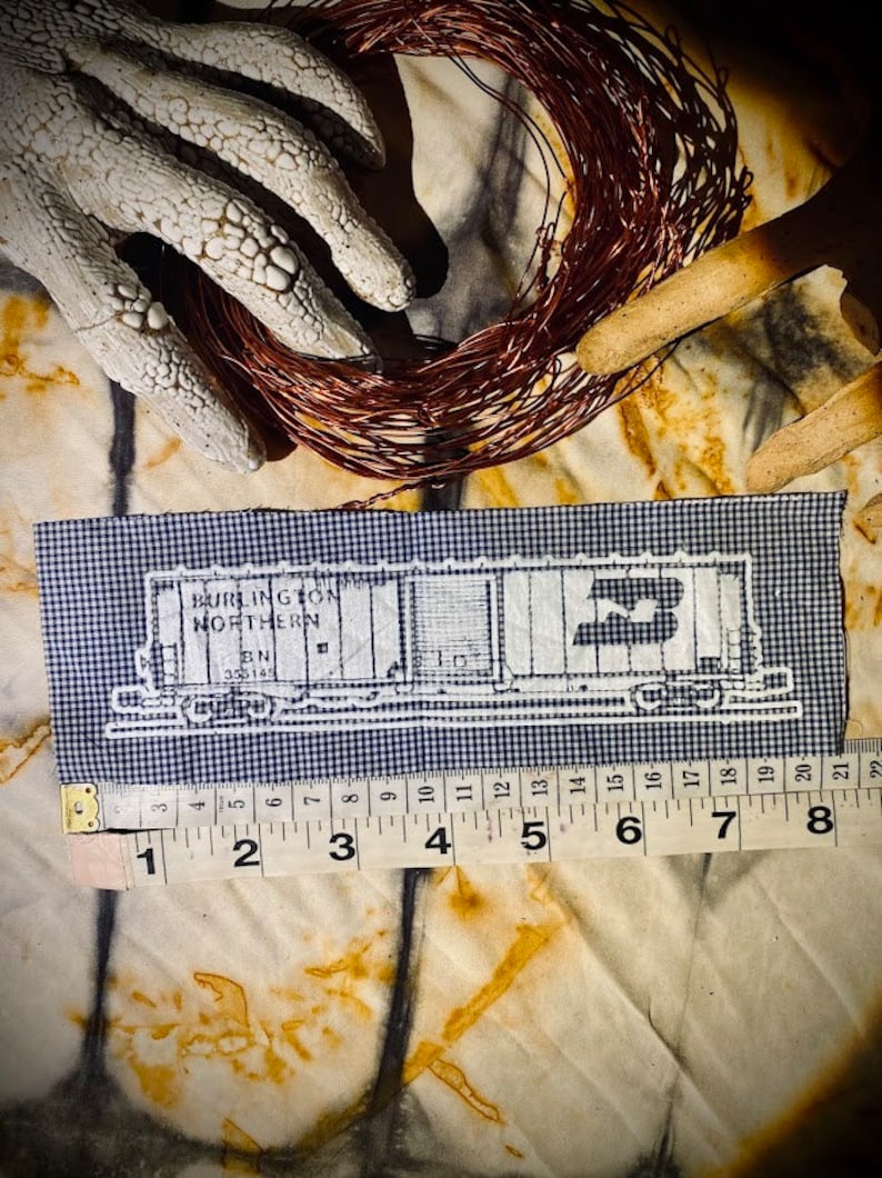 May include: A rectangular navy and white gingham fabric patch featuring a white embroidered outline of a Burlington Northern train car. A measuring tape is positioned along the bottom edge, showing a length of approximately 8 inches.