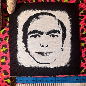 May include: A square black fabric patch featuring a high-contrast white screen-printed illustration of a man's face. A measuring tape placed along the bottom edge shows the patch width is approximately 4.25 inches wide.