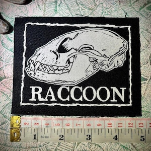 Puede incluir: Un parche rectangular de tela negra con una ilustración serigrafiada en blanco de un cráneo de mapache y la palabra RACCOON en letras mayúsculas. Una cinta métrica amarilla debajo muestra un ancho de unos 11,5 cm.