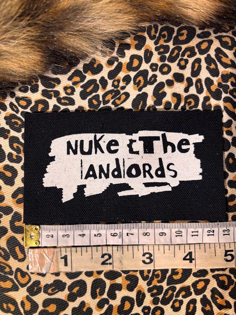 Peut inclure: &Eacute;cusson rectangulaire noir avec les mots "Nuke The Landlords" en blanc, lettrage vieilli. Un m&egrave;tre ruban indique que l'&eacute;cusson mesure environ 13 cm de long. Le fond est un motif l&eacute;opard.