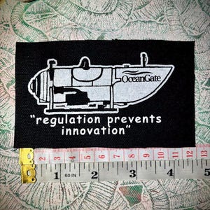 May include: Black and white patch with a drawing of a submersible with the text "OceanGate" on it. The text "regulation prevents innovation" is printed below the drawing. The patch is measured with a tape measure.