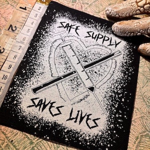 May include: Black and white patch featuring the text "SAFE SUPPLY SAVES LIVES" and a syringe design. The patch measures approximately 7.6 cm wide and 7.6 cm tall. The design is on a black background with a white splatter effect.