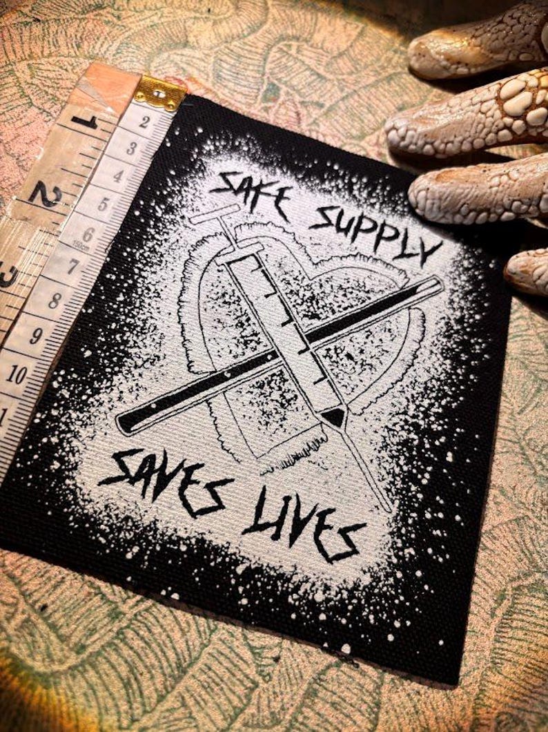 May include: Black and white patch featuring the text "SAFE SUPPLY SAVES LIVES" and a syringe design. The patch measures approximately 7.6 cm wide and 7.6 cm tall. The design is on a black background with a white splatter effect.