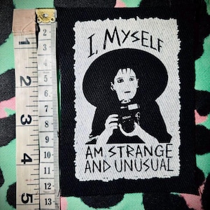 Könnte beinhalten: Schwarz-weißes Bügeleisen-Patch mit einer Person, die einen Hut trägt und eine Kamera hält. Der Text auf dem Patch lautet "I, Myself Am Strange And Unusual".