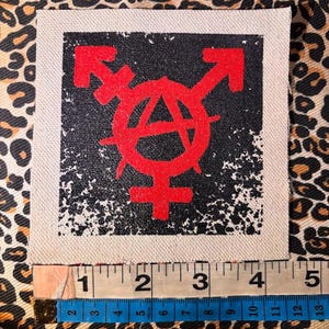 May include: A square fabric patch featuring a red graphic of a combined transgender and anarchy symbol on a black background. A measuring tape placed along the bottom edge shows the patch width is approximately 4.5 inches.