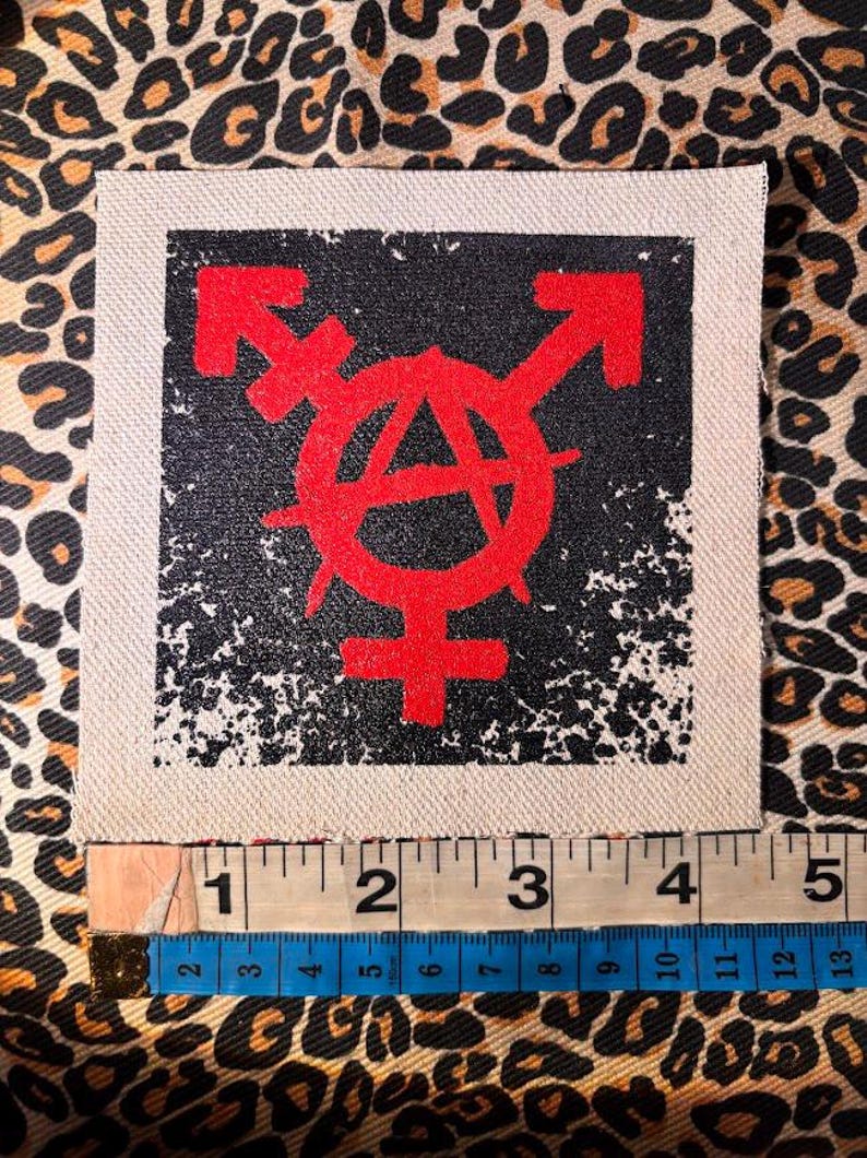 May include: A square fabric patch featuring a red graphic of a combined transgender and anarchy symbol on a black background. A measuring tape placed along the bottom edge shows the patch width is approximately 4.5 inches.