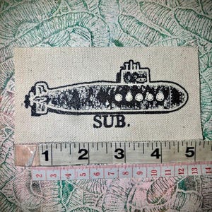 May include: A rectangular off-white fabric patch featuring a black screen-printed submarine graphic and the text SUB. A measuring tape is positioned below the patch, showing a width of approximately 12.7 cm for the printed design.