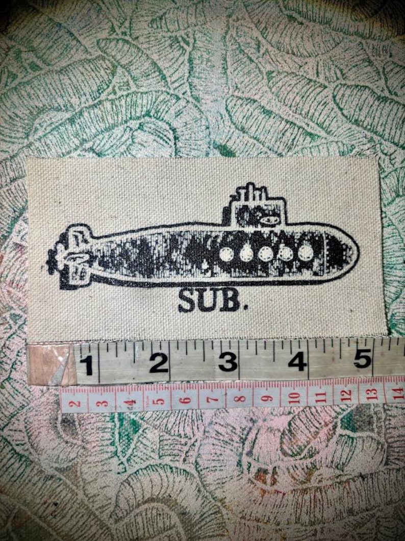 May include: A rectangular off-white fabric patch featuring a black screen-printed submarine graphic and the text SUB. A measuring tape is positioned below the patch, showing a width of approximately 12.7 cm for the printed design.