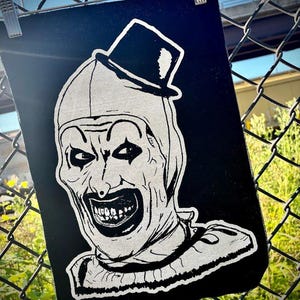 May include: A black and white graphic print of a scary clown wearing a top hat. The clown has a wide, toothy grin and is wearing a white hood and collar. The image is displayed on a chain-link fence.
