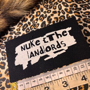 Peut inclure: &Eacute;cusson rectangulaire noir avec le texte "Nuke the Landlords" en blanc. Un m&egrave;tre ruban indique que l'&eacute;cusson mesure environ 10 cm de large. Le fond pr&eacute;sente un motif l&eacute;opard.